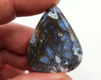 May include: A teardrop-shaped cabochon of blue, white, and gold agate with a swirling pattern. The agate has a natural, polished finish.