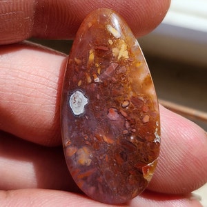 Könnte beinhalten: Ein tropfenförmiger Cabochon aus Achat mit einem braunen, orangen und weißen Muster. Der Stein hat eine glatte, polierte Oberfläche.