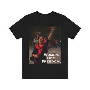 Peut inclure: T-shirt noir avec un graphisme d'une femme levant le poing, portant une écharpe rouge, et les mots "WOMEN. LIFE. FREEDOM." en blanc. L'arrière-plan est flou, montrant des personnes et une rue.