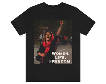 May include: Black t-shirt featuring a graphic of a woman with her fist raised, wearing a red scarf, and the words "WOMEN. LIFE. FREEDOM." in white. The image has a blurred background of people and a street.
