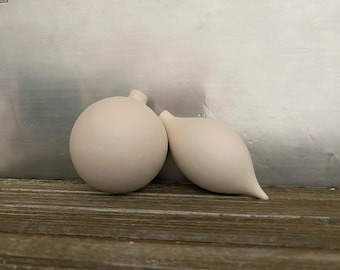 May include: Two white ceramic ornaments, one round and one teardrop shaped, on a wooden surface.