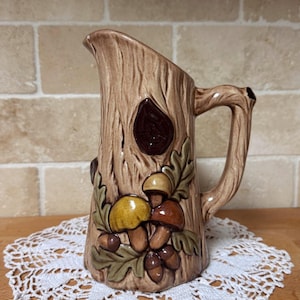 May include: A ceramic pitcher with a tree trunk design, featuring a handle and a spout. The pitcher is decorated with a cluster of mushrooms, acorns, and leaves in shades of brown, yellow, and green. It sits on a white lace doily.