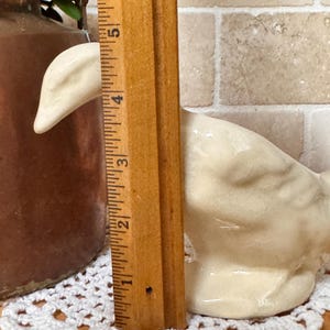 Op de afbeelding: Een cr&egrave;mekleurig keramieken ganzenbeeldje, ongeveer 10 cm hoog, staat op een wit gehaakt kleedje. Een houten liniaal is naast de gans geplaatst om te meten. De achtergrond omvat een bruine kruik en een bakstenen muur.