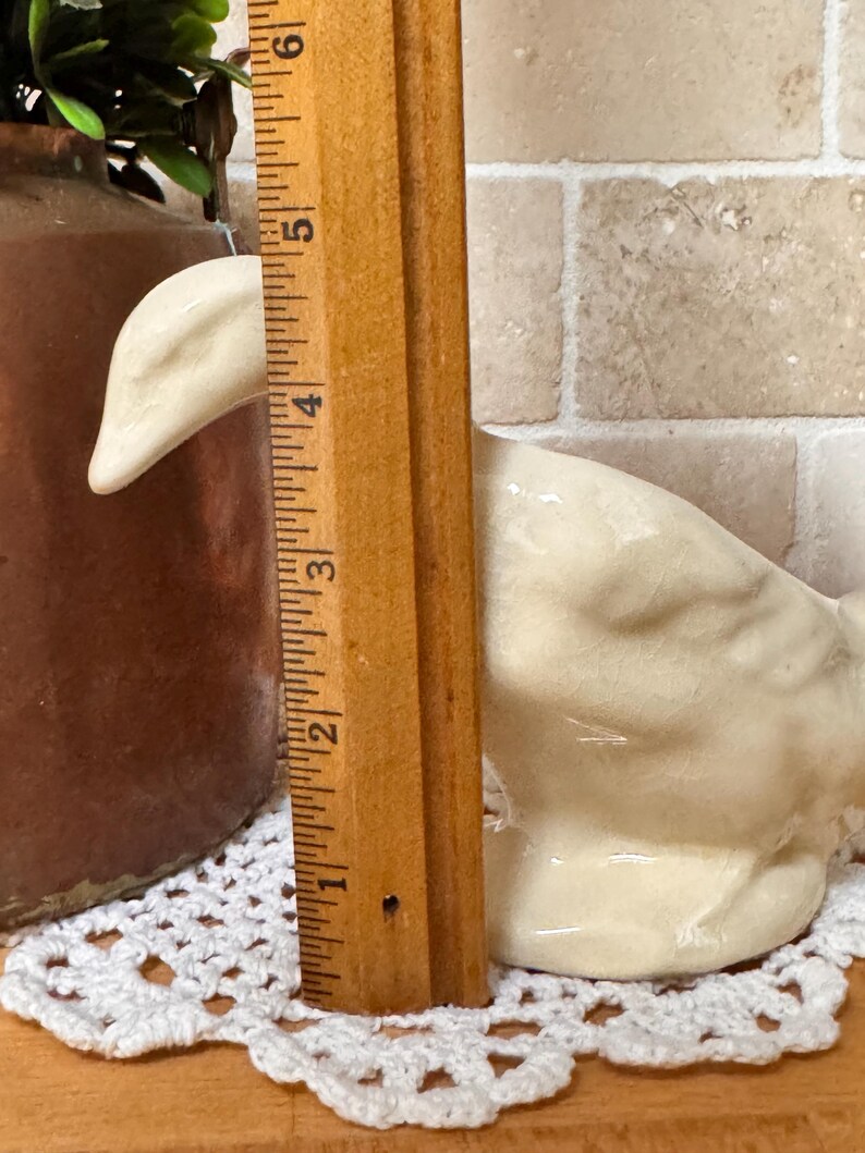 Op de afbeelding: Een cr&egrave;mekleurig keramieken ganzenbeeldje, ongeveer 10 cm hoog, staat op een wit gehaakt kleedje. Een houten liniaal is naast de gans geplaatst om te meten. De achtergrond omvat een bruine kruik en een bakstenen muur.