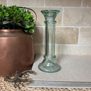 May include: A clear, light green glass candlestick holder with a flared base and top. The candlestick holder is cylindrical and sits on a neutral-colored surface. A copper pot with greenery and a bronze crayfish figurine are in the background.