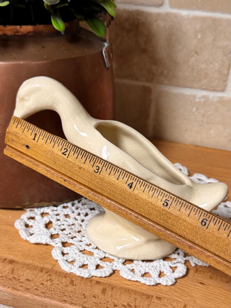 Op de afbeelding: Cr&egrave;mekleurige keramische eendvormige plantenbak, ongeveer 15 cm lang. De eend heeft een lange nek en een kleine basis. De plantenbak staat op een wit kanten kleedje. De achtergrond omvat een koperen container en een bakstenen muur.