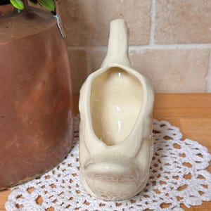 Op de afbeelding: Cr&egrave;mekleurige keramische eendvormige creamer met een brede opening en een handvat. De creamer heeft een gedetailleerde snavel en een getextureerd oppervlak. Het staat op een wit gehaakt kleedje, met een koperkleurige container en groen in de achtergrond.