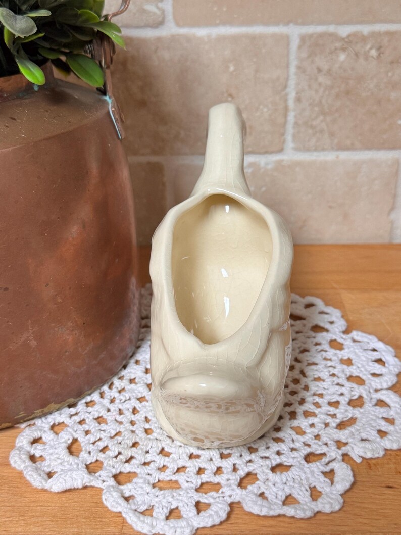 Op de afbeelding: Cr&egrave;mekleurige keramische eendvormige creamer met een brede opening en een handvat. De creamer heeft een gedetailleerde snavel en een getextureerd oppervlak. Het staat op een wit gehaakt kleedje, met een koperkleurige container en groen in de achtergrond.