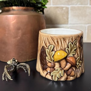 May include: A ceramic planter with a tree trunk design, featuring embossed mushrooms and acorns in shades of brown, yellow, and orange. The planter has a white interior and is suitable for small plants or decorative use.