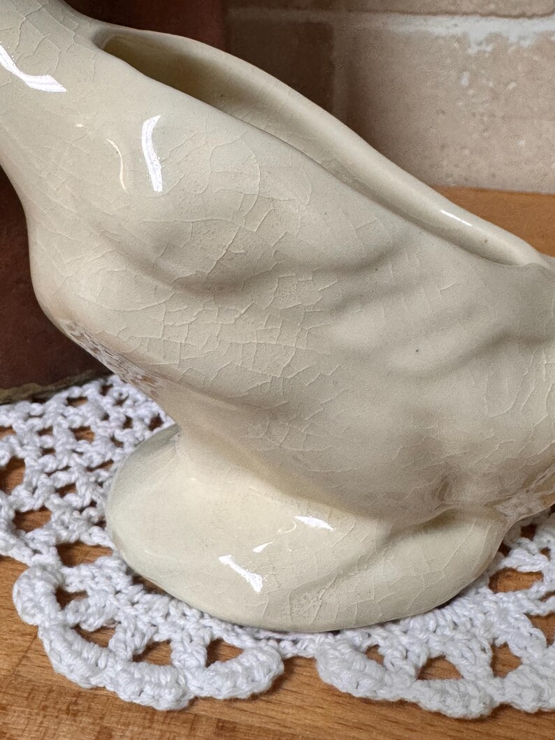 Op de afbeelding: Cr&egrave;mekleurig keramiek zwaanbeeldje met een getextureerd, gebarsten oppervlak. De zwaan heeft een gebogen nek en een brede basis. Het staat op een wit gehaakt kleedje, wat wijst op een vintage of antieke esthetiek.