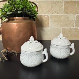 May include: Two white ceramic lidded jars with handles. The jars have a textured, dotted surface and a decorative finial on the lid. A copper pot with greenery and a bronze crab figurine are in the background.
