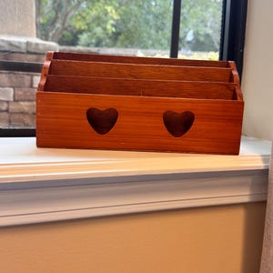 May include: A rectangular wooden letter holder with a medium brown finish. The front panel features two cut-out heart shapes. The organizer has three tiered slots for storing mail or documents, with a stepped design that increases in height toward the back.