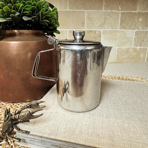 May include: A vintage, stainless steel coffee pot with a lid and handle. The pot is cylindrical with a spout for pouring. The background includes a copper container with greenery and a decorative bronze crab.