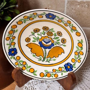 May include: A decorative ceramic plate with a floral design. The plate features a central arrangement of blue and yellow flowers, surrounded by a yellow border and a ring of orange dots. The plate is displayed on a wooden stand.