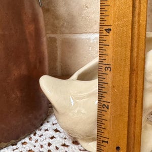 Op de afbeelding: Een cr&egrave;mekleurige keramische creamer met een tuit, ongeveer 5 cm hoog. De creamer heeft een craquel&eacute;-glazuur. Een houten liniaal staat ernaast voor de meting.