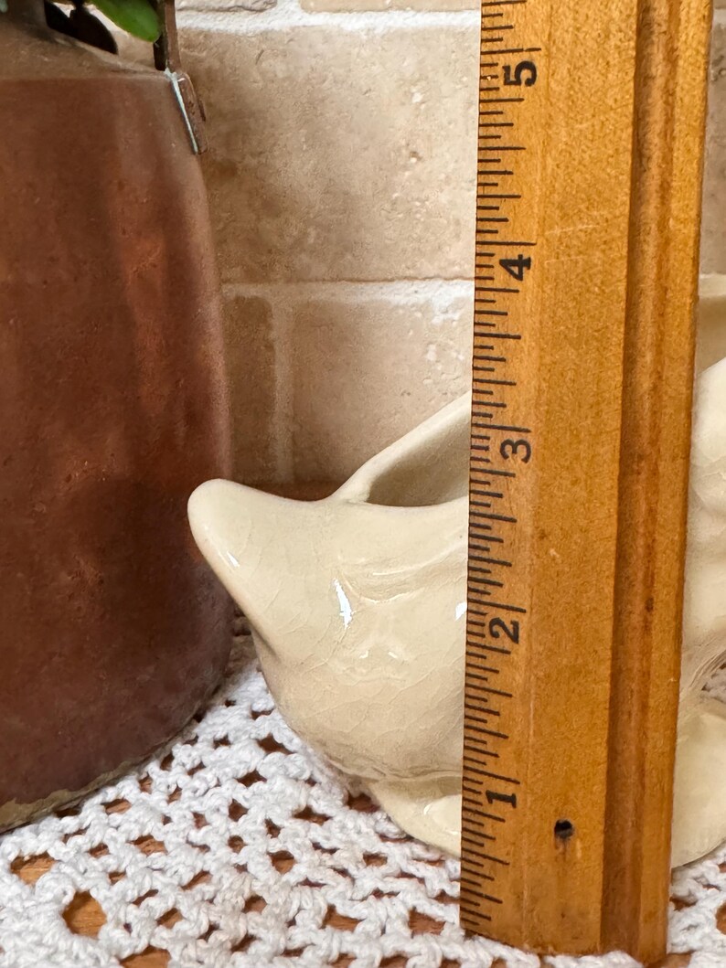 Op de afbeelding: Een cr&egrave;mekleurige keramische creamer met een tuit, ongeveer 5 cm hoog. De creamer heeft een craquel&eacute;-glazuur. Een houten liniaal staat ernaast voor de meting.