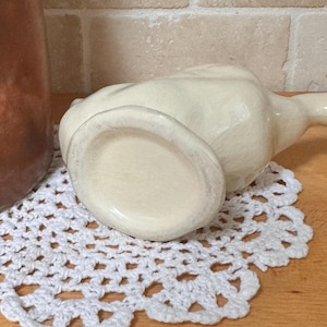 Op de afbeelding: Een cr&egrave;mekleurige keramische kan met een lange tuit, op een wit gehaakt kleedje. De kan heeft een gecraqueleerde glazuur en staat op een houten oppervlak. Een koperen container is gedeeltelijk zichtbaar op de achtergrond.