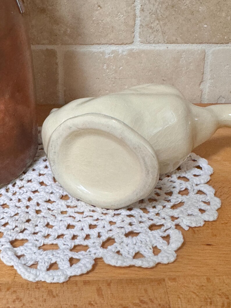 Op de afbeelding: Een cr&egrave;mekleurige keramische kan met een lange tuit, op een wit gehaakt kleedje. De kan heeft een gecraqueleerde glazuur en staat op een houten oppervlak. Een koperen container is gedeeltelijk zichtbaar op de achtergrond.