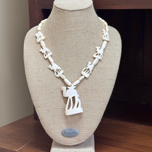 May include: Ivory-colored necklace featuring carved camel and animal figures. The necklace is displayed on a mannequin with a beige fabric. The necklace has a central camel pendant and smaller animal figures along the chain.