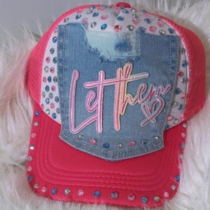 May include: A bright pink trucker hat featuring a white front panel adorned with scattered blue and pink rhinestones. A distressed denim patch is centered on the front, displaying the embroidered pink text Let them with a heart symbol.