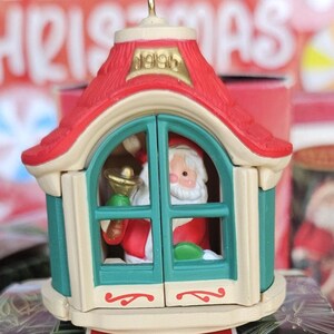 May include: A 1995 Hallmark Keepsake ornament featuring a miniature house with a red roof and teal walls. Inside the house, a Santa Claus figure holds a gold bell. The ornament sits atop its original box, which displays the text Hallmark KEEPSAKE ORNAMENT and FEATURES MOVEMENT.