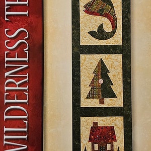 May include: A quilt pattern for a wall hanging called "Wilderness Trio" by Jeri Kelly. The pattern features a fish, a pine tree, and a cabin with a red roof. The pattern is 6 1/2 inches by 20 1/2 inches.