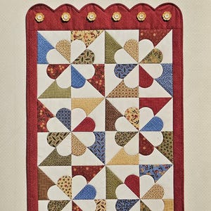 May include: A rectangular table runner with a red scalloped border and floral accents. The center features a patchwork design of white, red, blue, yellow, and green fabric in a pinwheel pattern. The text "Pinwheel Posies Table Runner" is at the top.