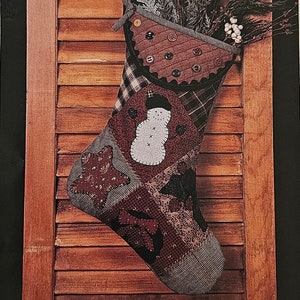May include: A patchwork Christmas stocking with a snowman design, hanging on a wooden door. The stocking is made of various fabrics in shades of brown, red, and black. The stocking is decorated with buttons and a black ribbon.