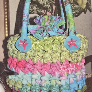 May include: A crocheted fabric bag with a green, pink, and blue color scheme. The bag has a fabric handle with a blue and purple floral pattern. The bag has two blue fabric circles with a red star on each.