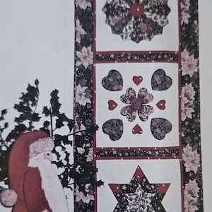 May include: A white quilted table runner with three different designs: a star, a flower, and a heart. The designs are made of black, red, and gold fabric. The runner is framed with a black and white floral border.