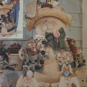 May include: A collection of handmade fabric bunny decorations. The largest bunny is wearing a straw hat with floral accents and a floral print jacket. The smaller bunnies are in a wicker basket and on a hay bale.