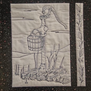 May include: A black and white embroidered image of a hand pump with a bucket on a wooden platform. The pump is surrounded by grass and a few blades of wheat. The image is framed with a black border and a vine border.