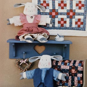 May include: Two handmade sheep dolls, one in a pink dress and the other in blue overalls, are displayed on a blue shelf with a heart cutout. A small quilt with red and blue squares hangs in the background, creating a rustic, country aesthetic.