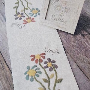 May include: White fabric with a scalloped edge, featuring an embroidered design of three colorful flowers with green stems and leaves. The text "Simple" and "Pretties" is embroidered in black cursive lettering.