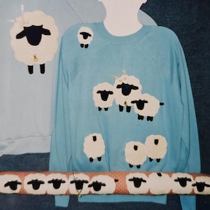 May include: Light blue sweatshirt with appliqued sheep design. The sheep are white with black faces and legs. The sweatshirt is displayed on a mannequin. A matching sheep-themed decorative item is in the foreground.