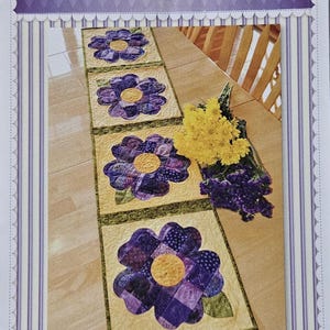 Op de afbeelding: Een patchwork tafelloper met grote paarse viooltjesbloemmotieven en gele centra op een lichtgele achtergrond. De loper is omrand met groen en heeft de tekst "Patchwork Pansy Table Runner". De bovenkant van de afbeelding heeft de tekst "Shabby Fabrics".