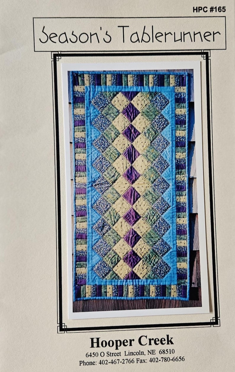 May include: A quilted table runner featuring a diamond pattern in blue, green, purple, and cream. The table runner has a blue border and the text "Season's Tablerunner" at the top.