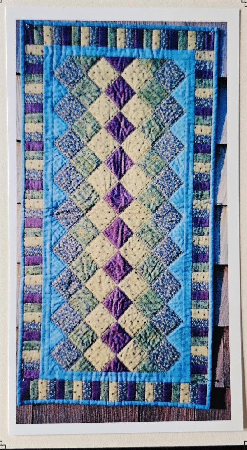 May include: A rectangular quilt with a diamond pattern in blue, purple, green, and cream. The quilt has a light blue border and a striped edge. The quilt is handmade and appears to be made of fabric. The dimensions are not visible.