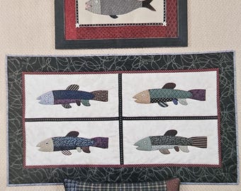 May include: A collection of fish-themed textile art pieces. Includes a framed picture of a fish with an American flag, a quilt with four fish, and a decorative pillow. The fish designs feature various patterns and colors.