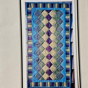 May include: A rectangular table runner with a patchwork design. The centre features a diamond pattern in shades of green, purple, and cream, framed by a blue border. The text "Season's Tablerunner" is at the top.