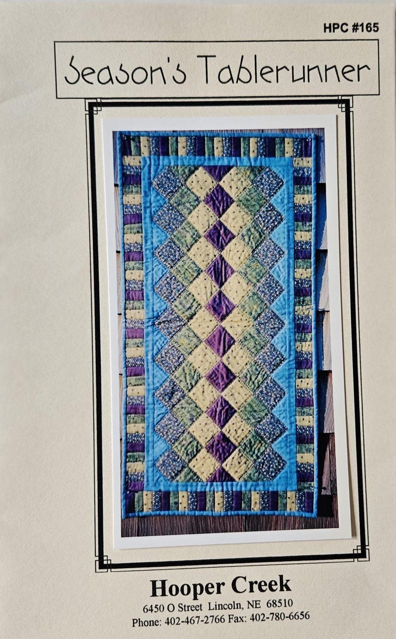 May include: A rectangular table runner with a patchwork design. The centre features a diamond pattern in shades of green, purple, and cream, framed by a blue border. The text "Season's Tablerunner" is at the top.