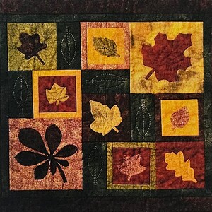 May include: A square fabric quilt featuring a patchwork design of autumn leaves in shades of deep red, burnt orange, mustard yellow, and dark green. The quilt displays various leaf shapes, including maple and chestnut, with some stitched outlines.