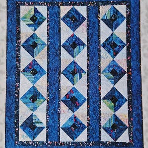 May include: A quilted textile featuring a repeating diamond pattern in shades of blue, white, and black. The quilt has a dark blue border and measures 60 inches by 72 inches.
