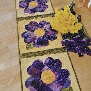 May include: A quilted table runner featuring four large purple flower appliqués with yellow centers on a light yellow background. The flowers have various patterned fabrics. Green trim and a bouquet of yellow flowers are also visible.