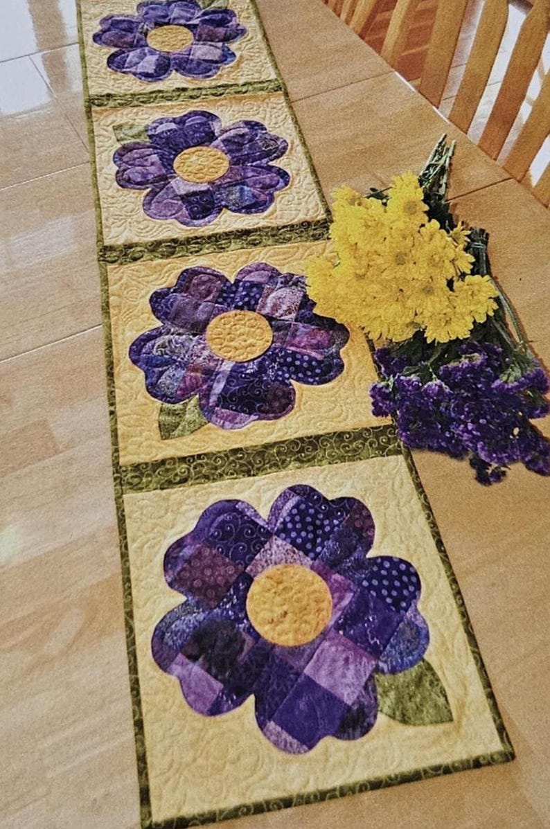 Op de afbeelding: Een gewatteerde tafelloper met vier grote paarse bloemenapplicaties met gele centra op een lichtgele achtergrond. De bloemen hebben verschillende patronen. Groene rand en een boeket gele bloemen zijn ook zichtbaar.