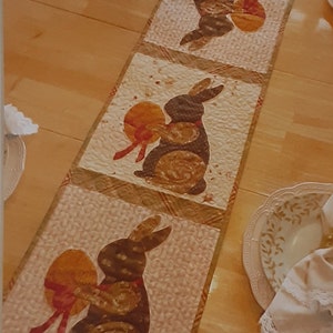 May include: A brown and white table runner with a quilted design featuring four brown bunnies holding orange eggs with red ribbons. The runner is on a wooden table with two plates and a napkin.