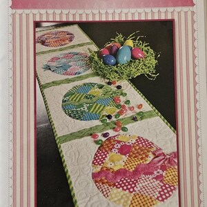 K&ouml;nnte beinhalten: Patchwork-Ostereier-Tischl&auml;ufer mit dekorativem Design. Der L&auml;ufer zeigt bunte, gemusterte Eierformen, ein Nest mit Eiern und den Text "Shabby Fabrics". Der L&auml;ufer ist etwa 100 cm lang.
