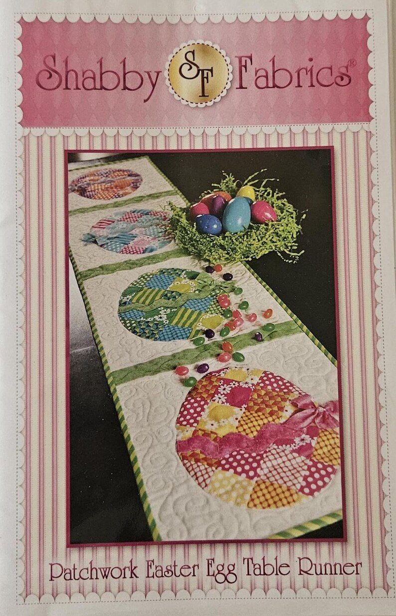 K&ouml;nnte beinhalten: Patchwork-Ostereier-Tischl&auml;ufer mit dekorativem Design. Der L&auml;ufer zeigt bunte, gemusterte Eierformen, ein Nest mit Eiern und den Text "Shabby Fabrics". Der L&auml;ufer ist etwa 100 cm lang.