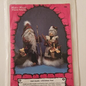 May include: A vintage craft pattern for making a Christmas Past Hob Knob doll set. The pattern includes instructions for assembling a Santa and Mrs. Claus doll with a small bear. The pattern is printed on pink paper with a white border and features a photo of the finished dolls.
