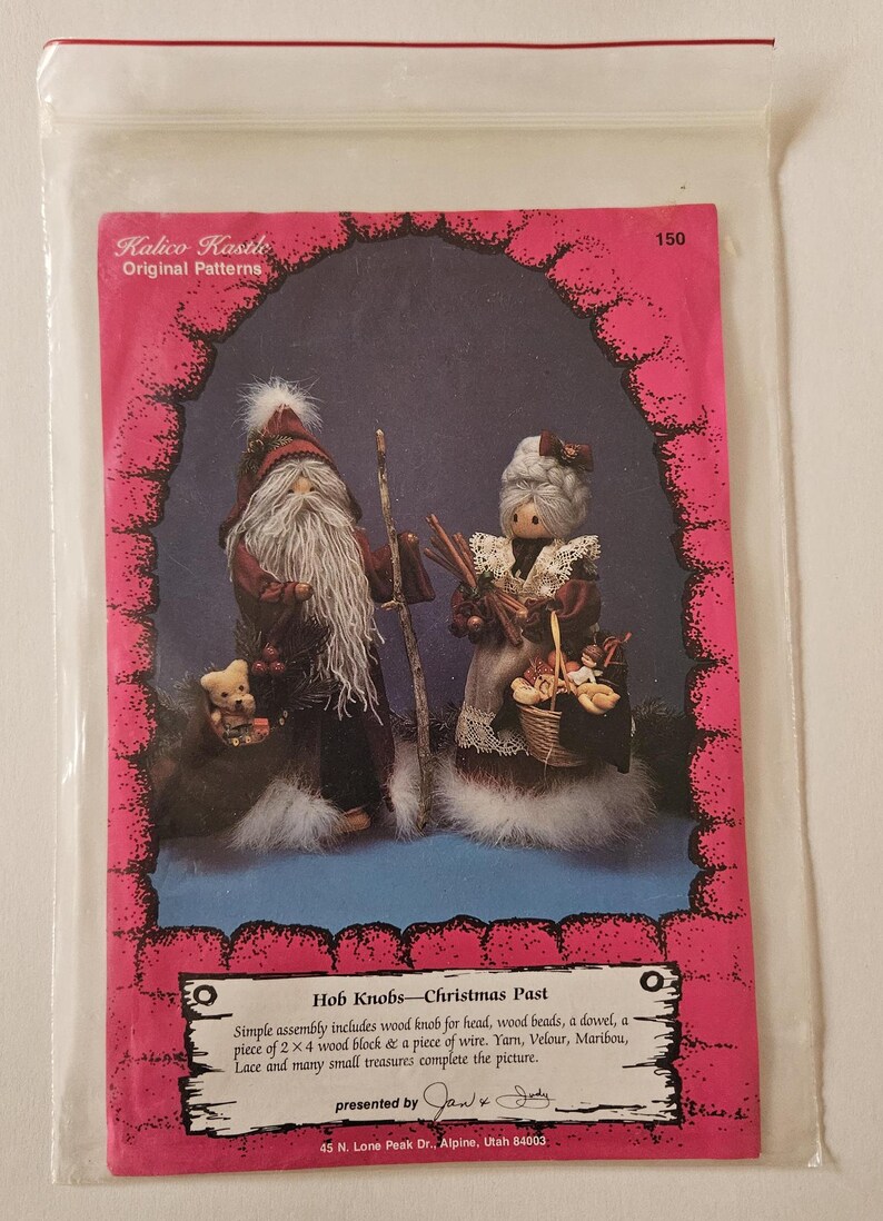 May include: A vintage craft pattern for making a Christmas Past Hob Knob doll set. The pattern includes instructions for assembling a Santa and Mrs. Claus doll with a small bear. The pattern is printed on pink paper with a white border and features a photo of the finished dolls.